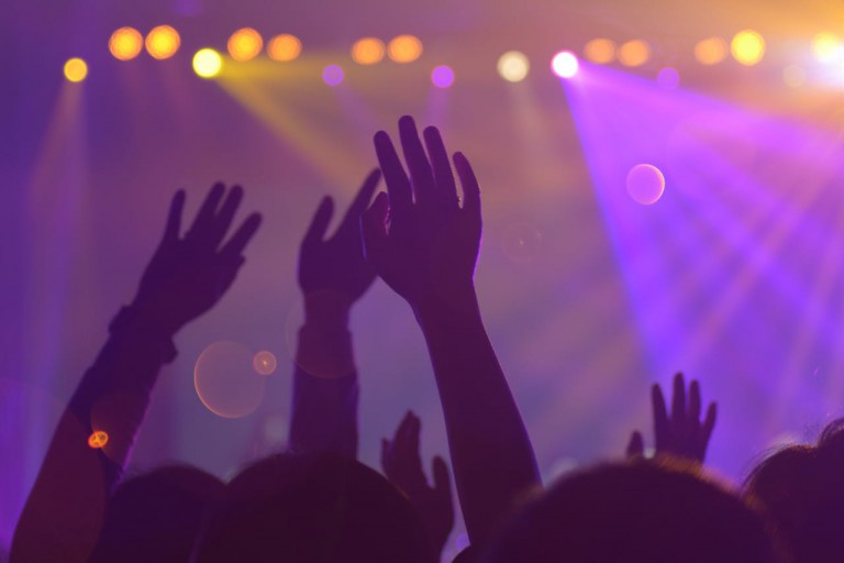 Audience with hands in the air at a night concert. Stage lights create orange / yellow bokeh in the purple hued background.