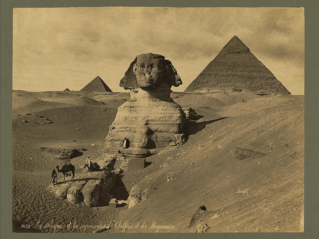 Men and camels atop the paw of the Sphinx, two pyramids in background.